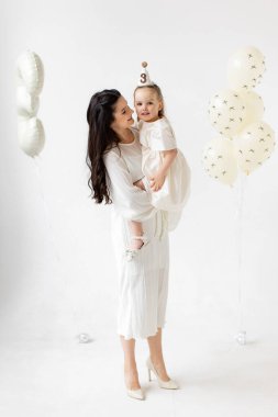 A mother holds her daughter, celebrating her third birthday with balloons and a party hat.