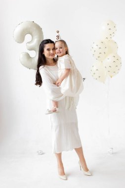 A mother holds her daughter, celebrating her third birthday with balloons and a party hat.
