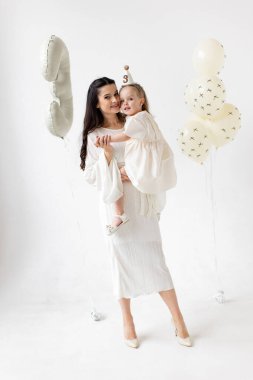 A mother holds her daughter, celebrating her third birthday with balloons and a party hat, all in white.