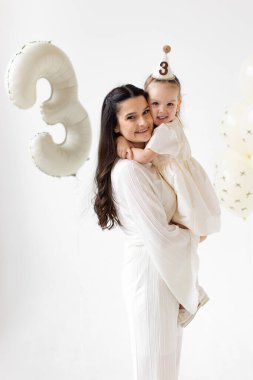 Bir anne, üçüncü doğum gününü balonlarla ve parti şapkasıyla kutluyor. İkisi de gülümsüyor.
