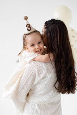 Bir anne, üçüncü doğum gününü kutlayan, doğum günü şapkası takan kızını kucaklar..