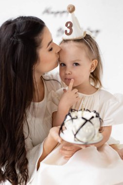 Bir anne, elinde pastayla kızının üçüncü doğum gününü kutlarken onu alnından öper..