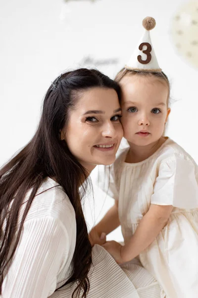 Bir anne ve küçük kızı kucaklaşıyorlar, kızlarının üçüncü doğum gününü kutlama şapkası ve gülümsemesiyle kutluyorlar..