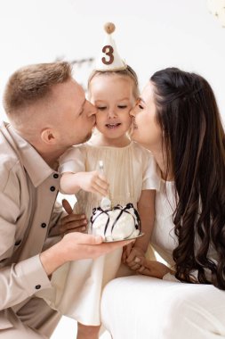 Genç bir kız üçüncü doğum gününü ailesiyle kutluyor. Pastasını tutarken yanaklarını öpüyorlar..
