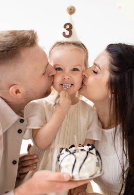 Genç bir kız, pasta yerken yanaklarını öpen anne ve babasıyla üçüncü doğum gününün tadını çıkarıyor..
