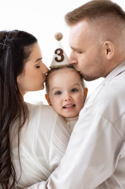 Sevgi dolu bir aile üçüncü yaş gününü öpücüklerle kutlar. Ebeveynler parti şapkası takan kızlarını kucaklıyor..