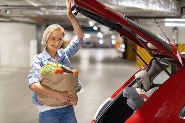 Gülümseyen bir kadın, bir yeraltı otoparkındaki arabasının bagajına yiyecek dolduruyor.