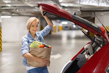 Gülümseyen bir kadın bir otoparktaki arabasının bagajına yiyecek dolduruyor.