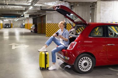 Kırmızı bir arabanın açık bagajında oturan bir kadın, otoparkta beklerken telefonunu kullanıyor.