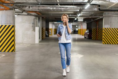 Bir kadın elinde kahve fincanı ve telefonla konuşurken otoparkta yürür.