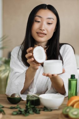 Beyaz cüppeli genç bir kadın, avokado ve nane gibi doğal malzemelerle çevrili yarım hindistan cevizi ve bir krema kavanozu tutuyor.