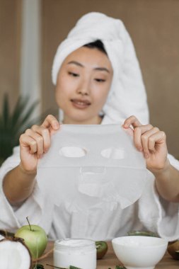 Kafasına havlu sarılmış genç bir kadın, içinde kivi, elma ve hindistan cevizi gibi malzemeler bulunan bir çarşaf maskesi tutuyor.