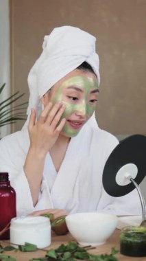 Kafasına havlu sarılmış genç bir kadın, yeşil bir yüz maskesi takar. Aynaya bakar, cilt bakımı malzemeleriyle çevrilidir.
