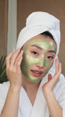 Kafasına havlu sarılmış genç bir kadın beyaz bornoz giyerken yeşil yüz maskesi takıyor.