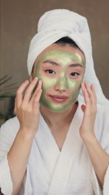 Kafasında havlu ve beyaz bornoz olan genç bir kadın, doğal cilt bakımı için gerekli malzemelerle çevriliyken yeşil yüz maskesi takıyor.
