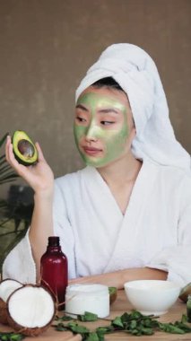 Kafasında havlu ve beyaz bornoz olan genç bir kadın avokado maskesi takarken elinde yarım bir avokado tutuyor.