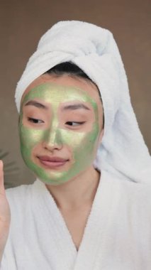 Kafasında havlu ve beyaz bornoz olan genç bir kadın elinde elma ve bir kavanoz ürün tutarken yeşil yüz maskesi takıyor.