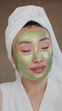 Kafasında beyaz bir havlu ve beyaz bir bornoz olan genç bir kadın elinde elma ve krema kavanozu tutarken yeşil bir maske takıyor.