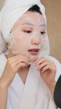 Kafasında havlu olan genç bir kadın, beyaz bir bornoz giyerken yüzüne hidratasyon maskesi takıyor ve kaplıcaya hazırlanıyor.