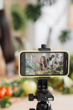 Genç bir kadın, arka planda taze sebzeler ve bitkilerle sağlıklı bir yemek hazırlarken kendini filme çekiyor.