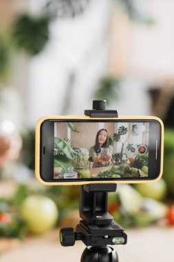 Genç bir kadın akıllı telefonunda bitkilerle çevrili sağlıklı beslenme ve taze malzemelerle ilgili bir video kaydediyor.