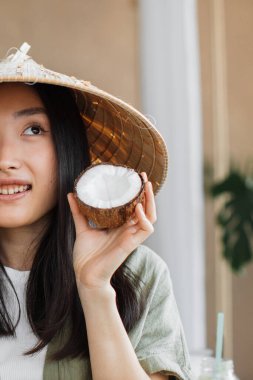 Konik şapka takan genç bir Asyalı kadın gülümserken yarım hindistan cevizi tutuyor.