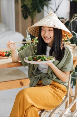 Konik şapka takan genç bir Asyalı kadın, yeşilliklerle çevrili, ahşap bir masada yemek çubuklarıyla sağlıklı bir salatanın tadını çıkarıyor.