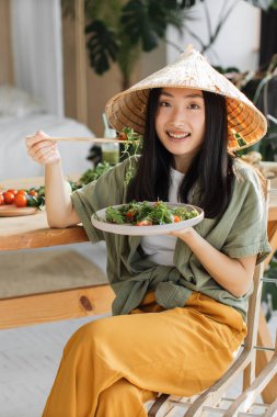 Konik şapka takan genç bir Asyalı kadın ahşap masada çubuklarla taze salata yerken gülümsüyor.