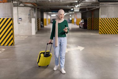 Başörtüsü takan bir kadın, otoparktan geçerken telefonuna bakar ve bavulunu çeker.