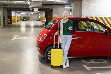 Garajda tesettürlü bir kadın arabasının yanında duruyor ve telefonuna bakıyor.