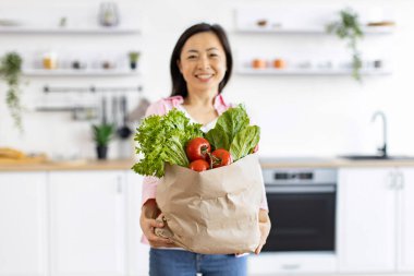 Gülümseyen bir kadın, parlak ve modern mutfağında içi taze ürünlerle dolu bir kesekağıdı tutuyor.