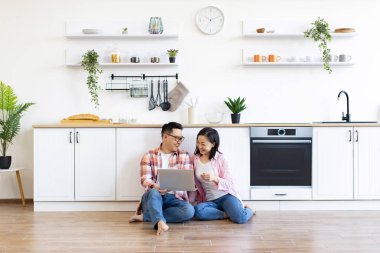 Gülümseyen bir çift modern mutfaklarında dizüstü bilgisayar kullanarak yerde oturuyor.