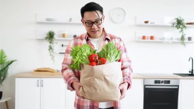 Asyalı bir adam, parlak, modern mutfağında içi taze ürünlerle dolu bir kesekağıdı tutarken gülümsüyor.