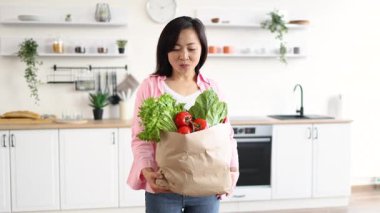 Asyalı bir kadın, parlak ve modern mutfağında elinde taze ürünlerle gülümsüyor.