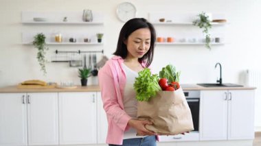 Gülümseyen bir kadın, parlak, modern mutfağında bir torba taze meyve tutar.