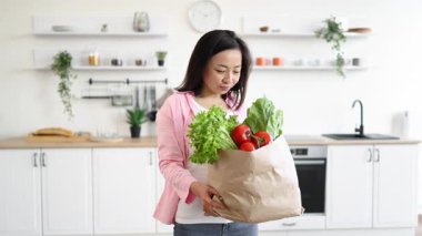 Gülümseyen bir kadın, parlak ve modern mutfağında içi taze ürünlerle dolu bir kesekağıdı tutuyor.