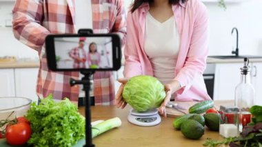 Bir çift, iyi aydınlatılmış bir mutfaktaki ahşap bir masada taze sebzelerle bir yemek videosu hazırlıyor.
