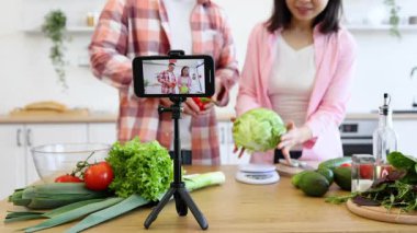 Bir çift, taze sebzeler ve tripodun üzerinde akıllı bir telefonla çevrili mutfağında bir pişirme videosu kaydediyor.