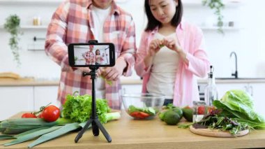 Asyalı bir çift mutfağında sağlıklı bir yemek hazırlıyor ve çevrimiçi izleyiciler için yemek pişirme dersi hazırlıyor.