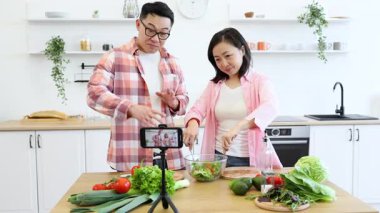 Asyalı bir çift modern mutfaklarında salata hazırlıyor, çevrimiçi seyircileri için bir yemek videosu çekiyor.