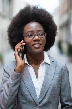 Afro saçlı profesyonel bir siyahi kadın kulağına akıllı telefon dayayıp konuşurken gözlük takıyor ve gri takım elbise giyiyor.