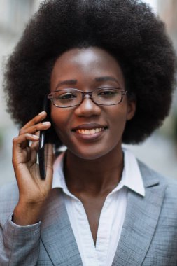 Uzun saçlı, profesyonel bir zenci kadın dışarıda akıllı telefonuyla konuşurken gözlük takıyor ve takım elbise giyiyor.