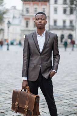 Takım elbiseli genç siyahi bir adam kaldırım taşı sokağında elinde deri bir çantayla dikiliyor.