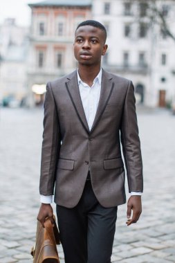 Takım elbiseli genç siyahi bir adam kaldırım taşlı bir sokakta yürür, elinde deri bir çantayla, düşünceli bir şekilde uzaklara bakar.