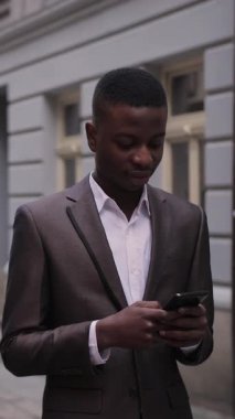 Kendine güvenen Afro-Amerikan işadamı şık takım elbiseli, caddede duruyor ve akıllı telefon kullanıyor. Modern cihazların yardımıyla internet üzerinden sorunları çözen yakışıklı bir adam..