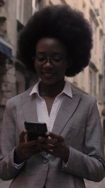 Zarif bir iş elbisesi içindeki Afro-Amerikalı kadın akıllı telefonuyla internette sörf yaparken şehir sokaklarında dikiliyor. Gözlüklü çekici bayan elinde modern aletlerle dışarıda poz veriyor..