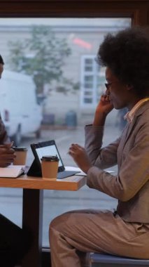 Afro-amerikan iş adamlarının resmi kıyafetleri içinde kafeteryada oturup kahve içerken, modern aletler kullanırken ve iletişim kurarken yan görüntüsü. İşbirliği ve çalışma toplantısı kavramı.