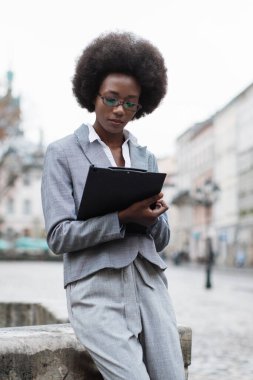 Afro saçlı, gözlüklü, takım elbiseli, şehir merkezinde elinde bir pano tutan profesyonel bir siyahi kadın.