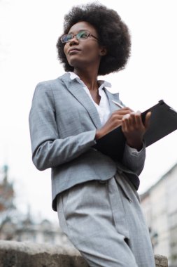 Takım elbiseli ve gözlüklü profesyonel bir siyahi kadın kameradan kafasını çevirirken düşünceli bir şekilde panoya yazı yazıyor.