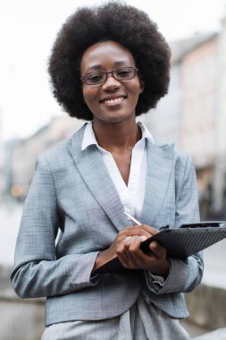 Afro saç stili ve gözlüklü profesyonel bir siyahi kadın, elinde bir pano ve kalem tutarken gülümsüyor ve iş ya da akademik bir ortam öneriyor.